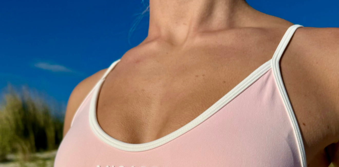 Close-up of a woman wearing the ANCARTA Vital Set in a soft pink color with white trim. The seamless sports bra features the ANCARTA logo on the chest. The background shows soft white sand and beach grass under a clear blue sky. Bold white text with a black outline across the image reads “VITAL SET.” This image is used as a thumbnail for a video in a welcome discount email.