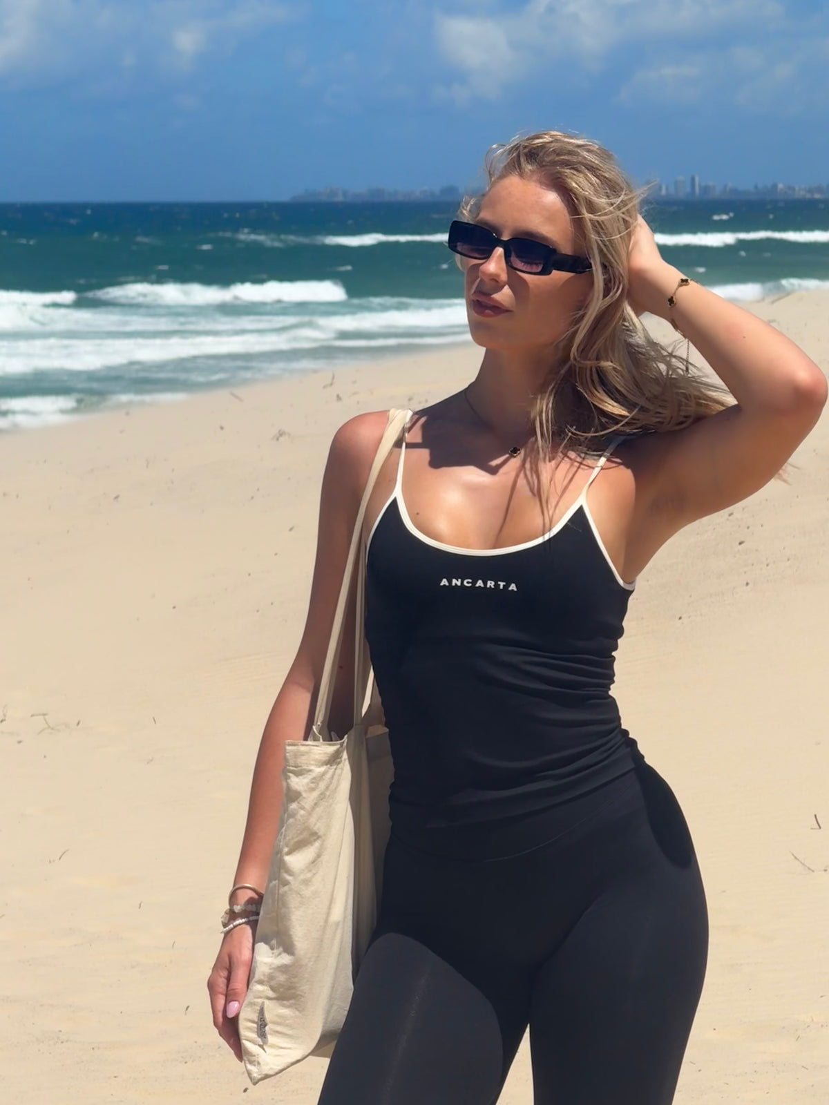 Woman on the beach in a black Ancarta tank and leggings with a canvas tote