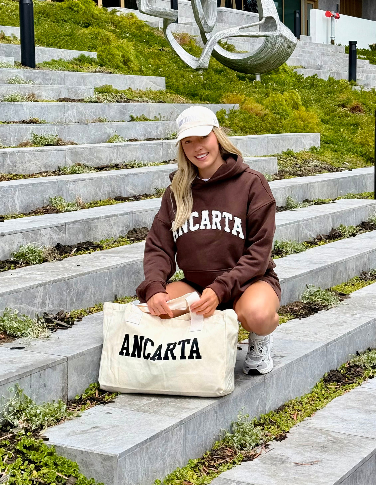 Smiling woman in the Ancarta Organic Cotton Relaxed Hoodie in Chocolate, with an Ancarta tote bag, on outdoor steps